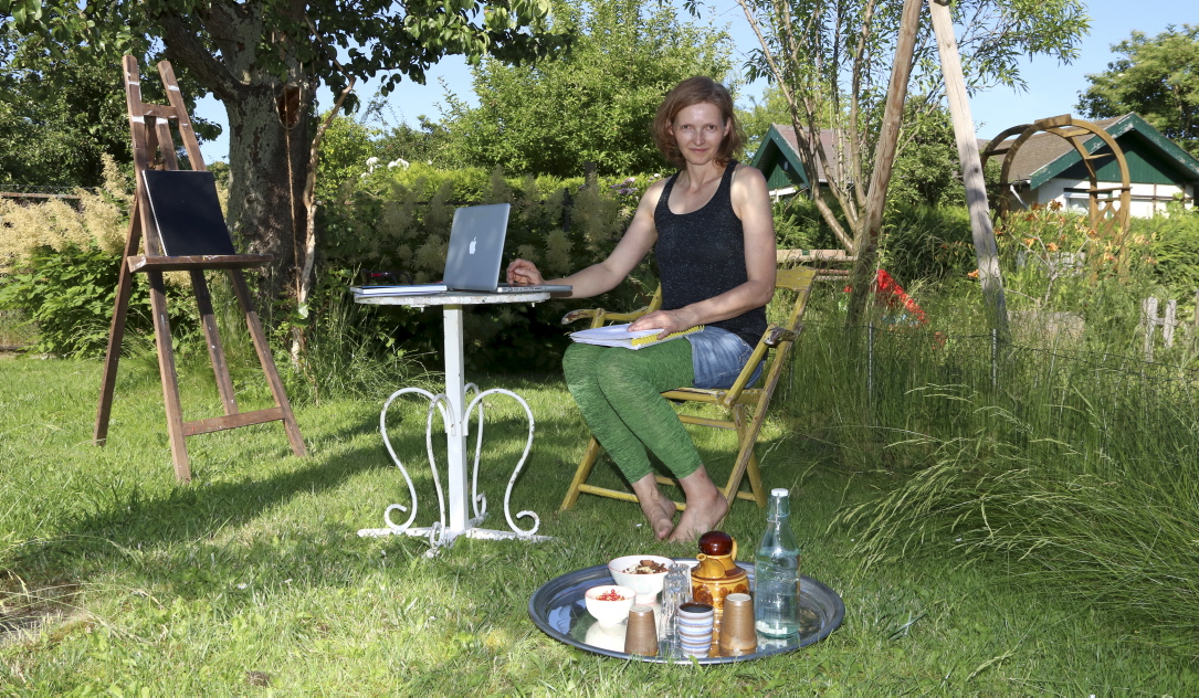 Die Künstlerin Claudia Michaela Kochsmeier in ihrem Gartenatelier. Sie sitzt auf der Wiese mit Laptop, Schreibzeug und einem großen Tablett mit Wasserkaraffe, Gartenkräutertee und frischen Früchten. Im Hintergrund ein alter Birnbaum und eine Staffelei mit einem großen Skizzenbuch.